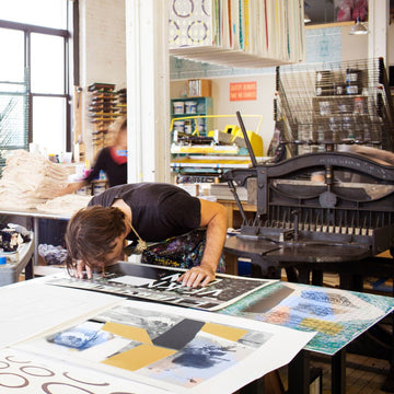 An artist leaning over a large art print in a busy printmaking studio