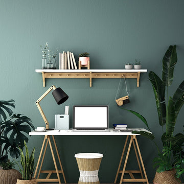 A stylish home study room with a white trestle desk, wooden lamp, laptop, wall shelf, and lush green plants