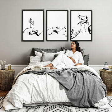 South Asian woman in white linen pyjamas lying in bed looking up at three black and white abstract wall art prints in a cosy Scandi bedroom