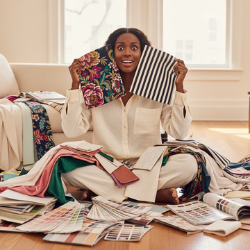Black woman sitting on floor surrounded by fabric swatches and decor samples, holding two swatches to her cheeks with a wide-eyed baffled grin, confused about which to choose for her interior design project