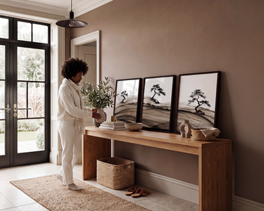 A woman in a white robe stands beside a wooden console table in a styled entryway, holding a plant. Three framed black-and-white landscape prints lean against a taupe wall, with decorative objects on the table and a woven basket underneath.
